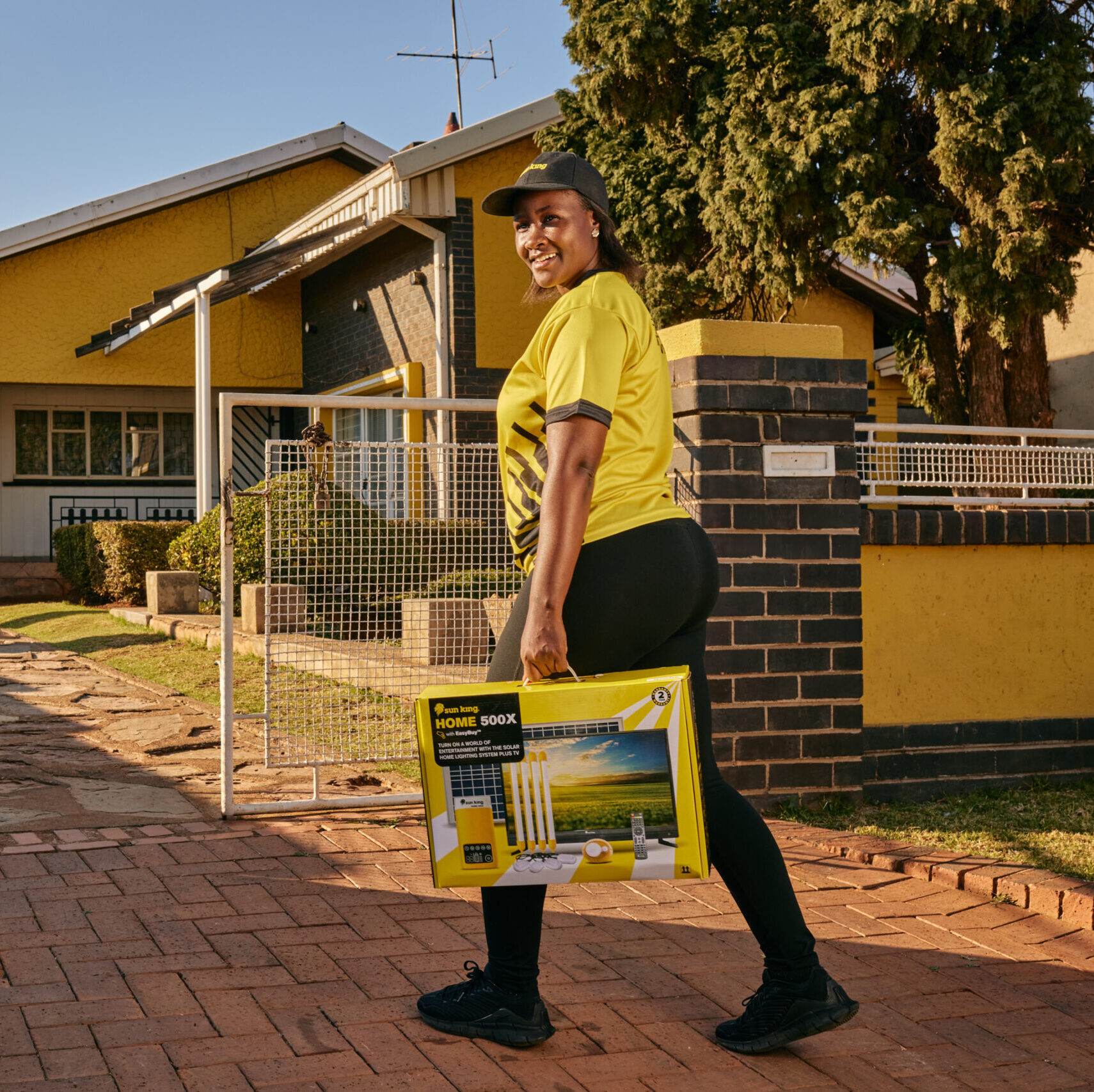 Sun King employee walks through a gate to a customer's house, carrying a Sun King Home 500X home solar powered lighting system complete with solar panels.