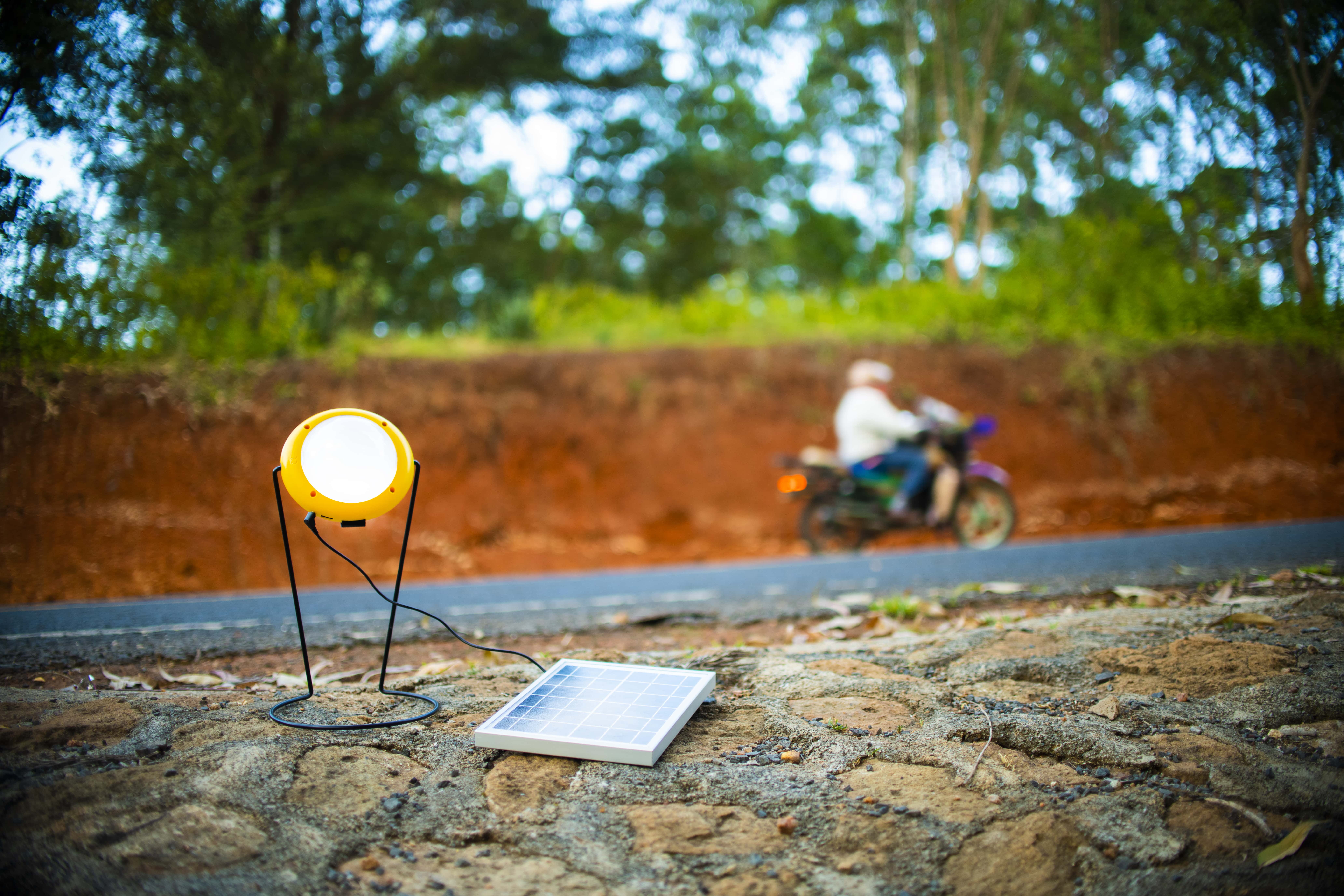A Sun King Pro 400 sits on the site of a road, while a man on a motorbike rides past. The Pro 400 portable, long-lasting solar powered light is plugged into a solar panel.