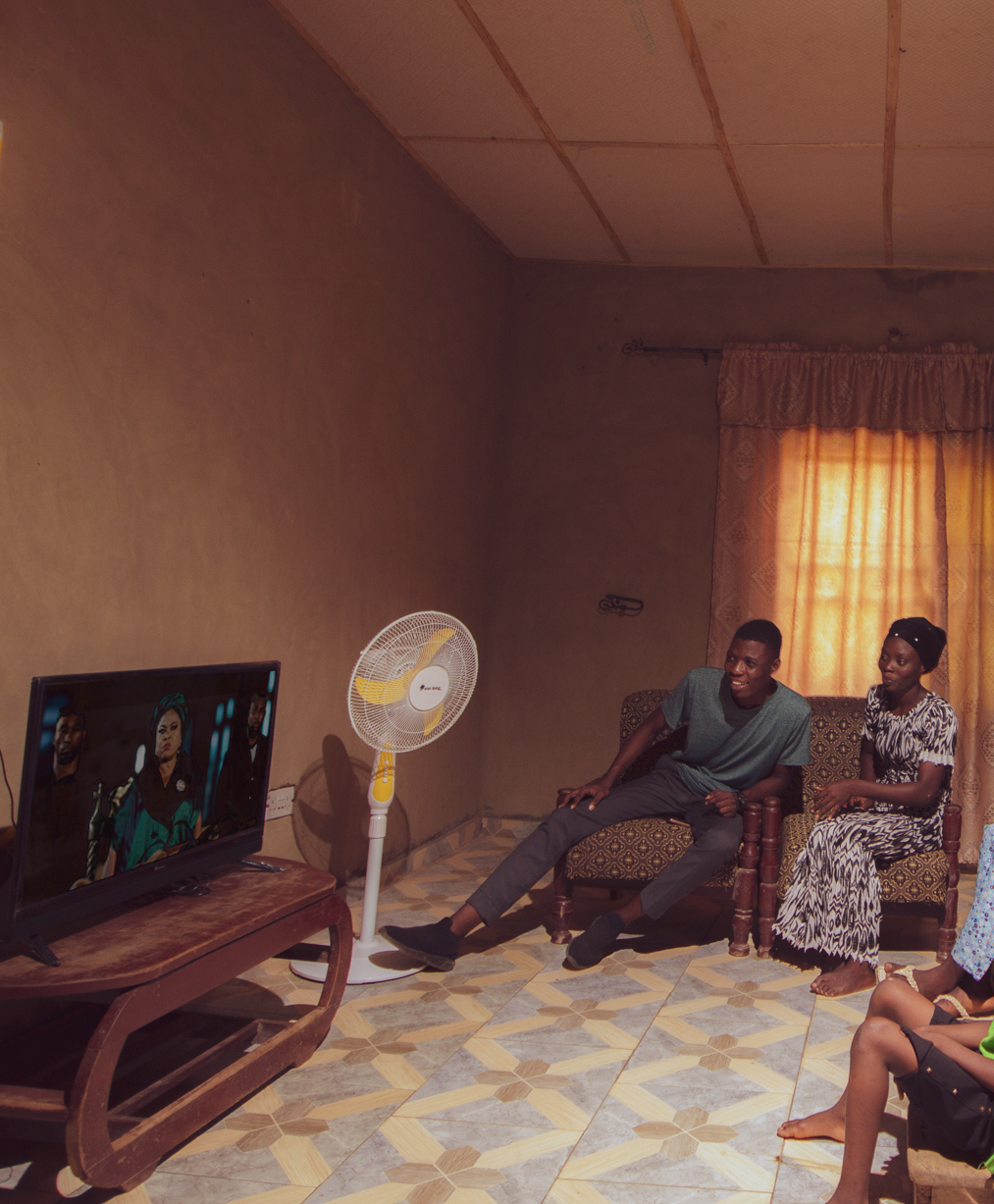A family sits around their living room which is lit by their Sun King Home 500X tube lights, and cooled by their Sun King pedestal fan.