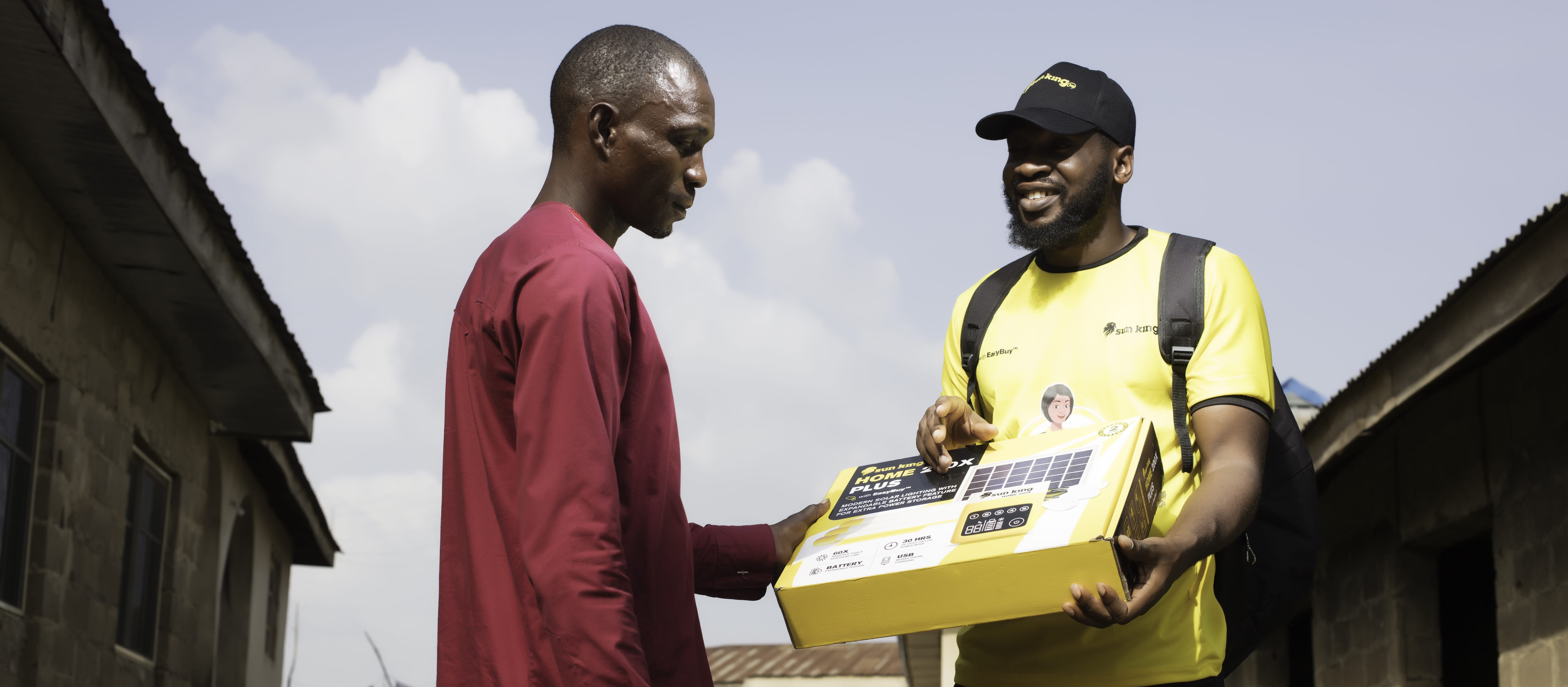 A Sun King field sales agent dressed in yellow stands next to a customer, who is dressed in red. The field sales agent is holding a Sun King product in a box.